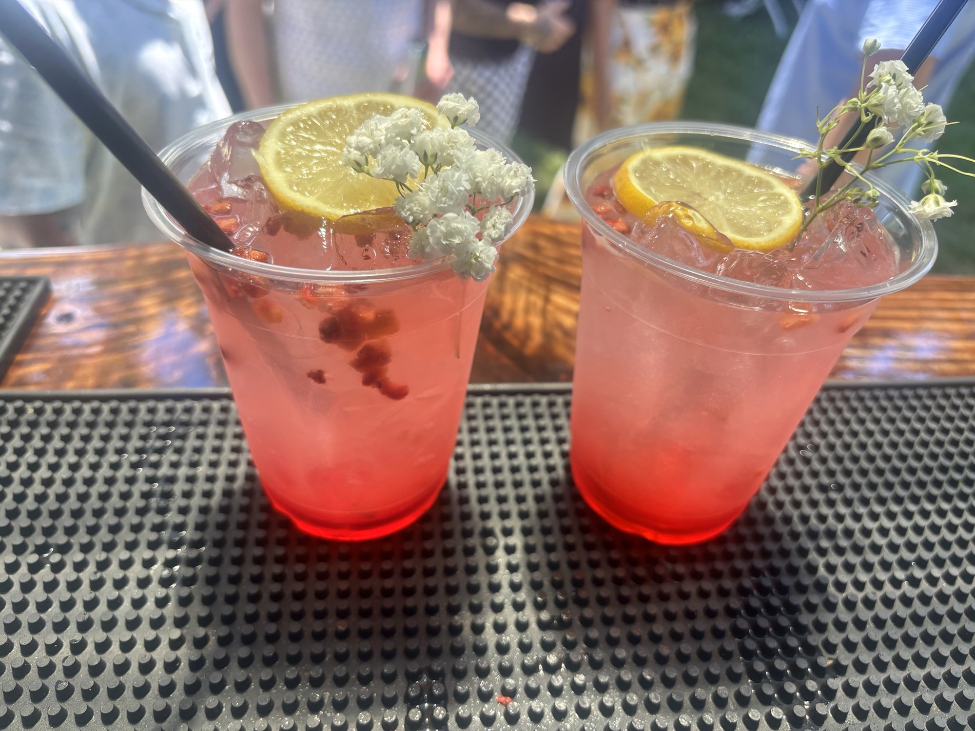 Two pink iced drinks in clear cups with lemon slices, small white flowers, and black straws on a bar mat