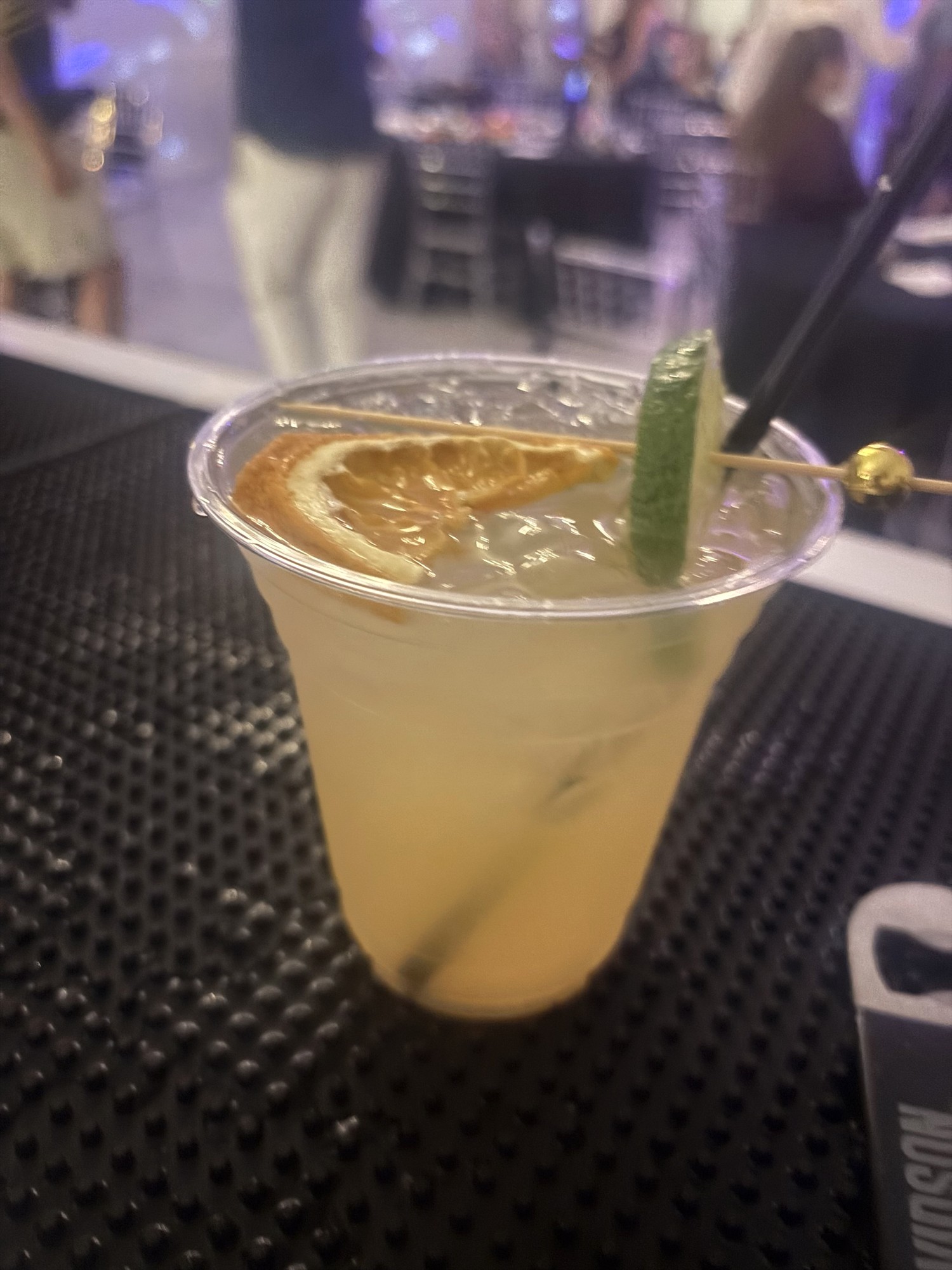 Pale yellow iced drink with an orange slice, a skewered lime slice, and a black straw
