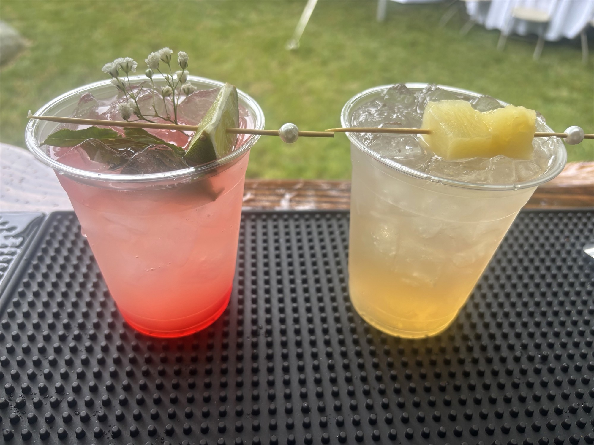 Pink and pale yellow iced drinks with lime and pineapple garnishes in clear cups on a bar mat