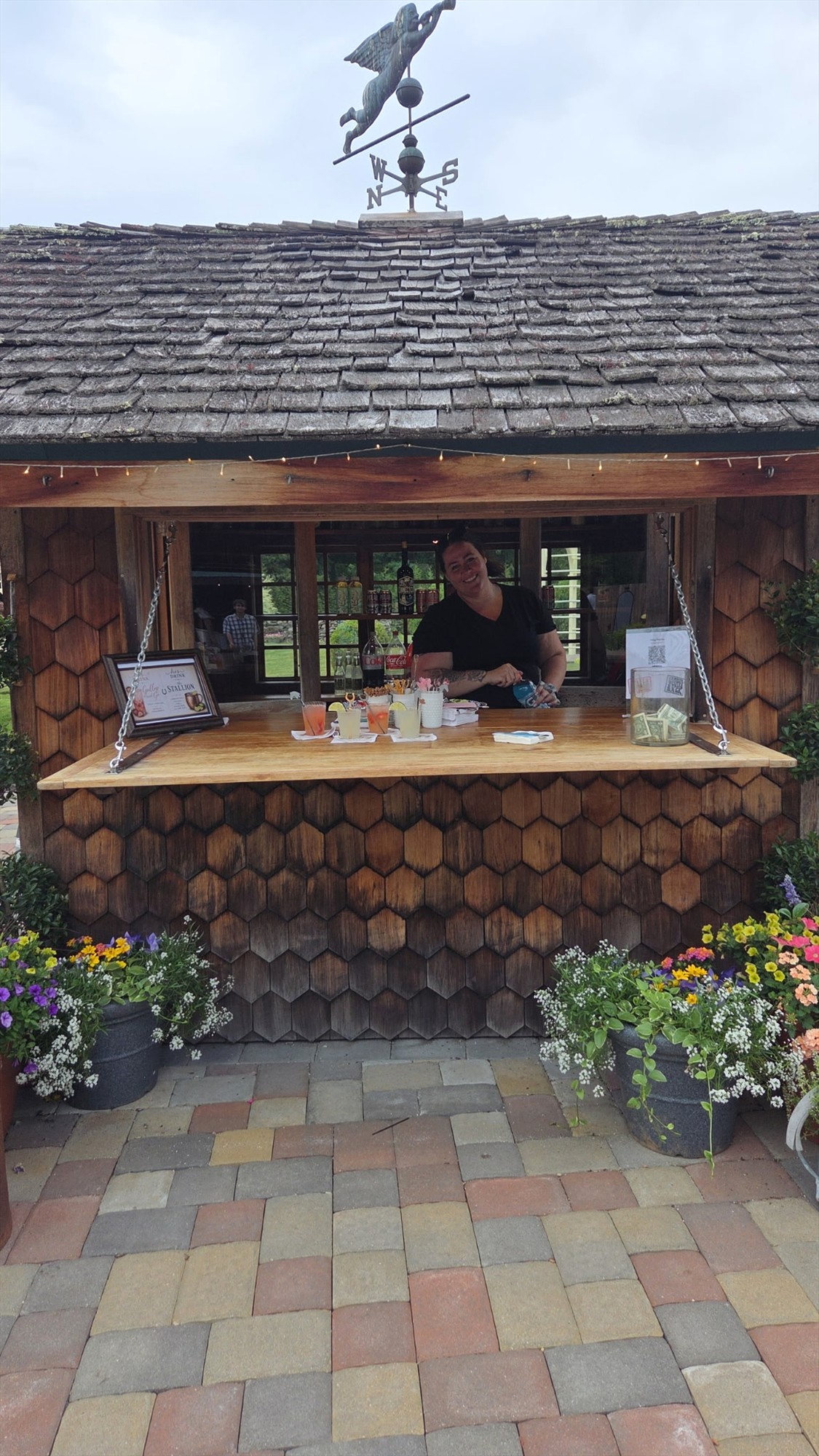 Person behind a wooden shingle bar with drinks on the counter and a horse weathervane above the roof