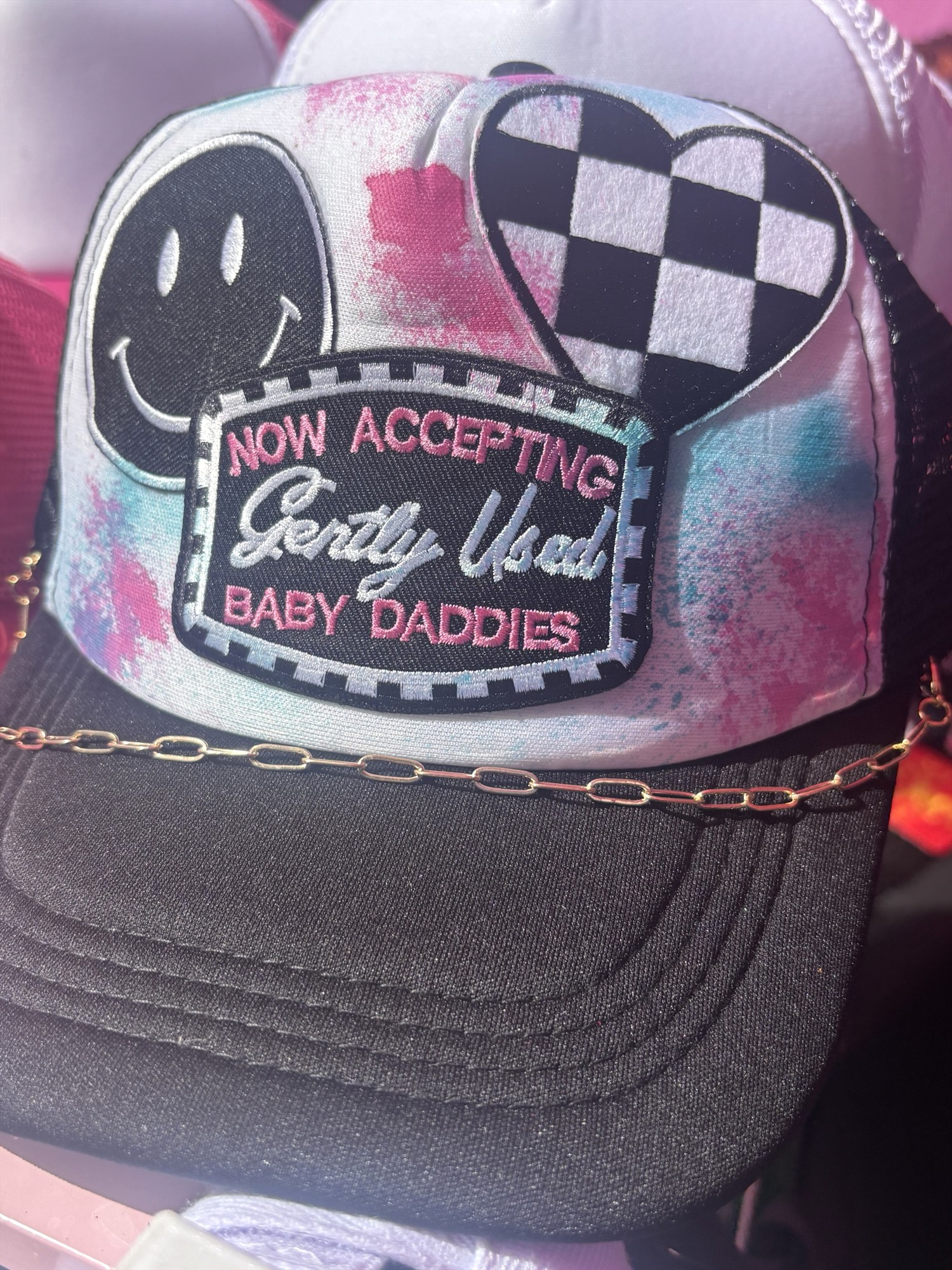 Black trucker hat with a pink and blue front, smiley face, checkered heart, Baby Daddies slogan patch, and gold-colored chain