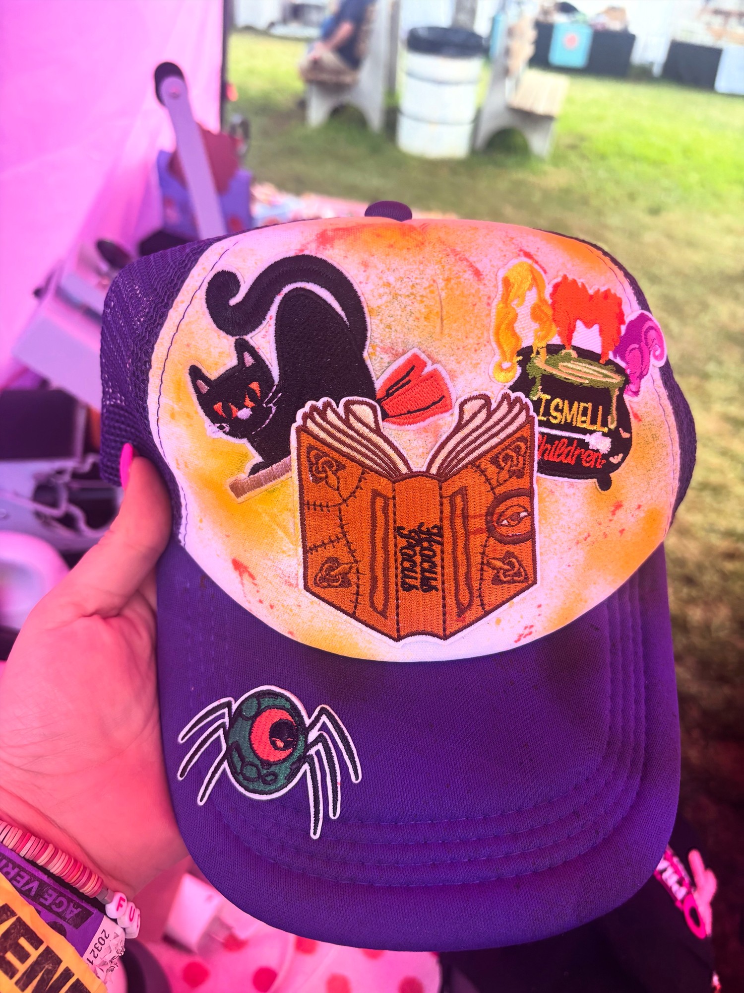 Hand holding a purple trucker hat with an orange and yellow front and black cat, spellbook, cauldron, and spider patches