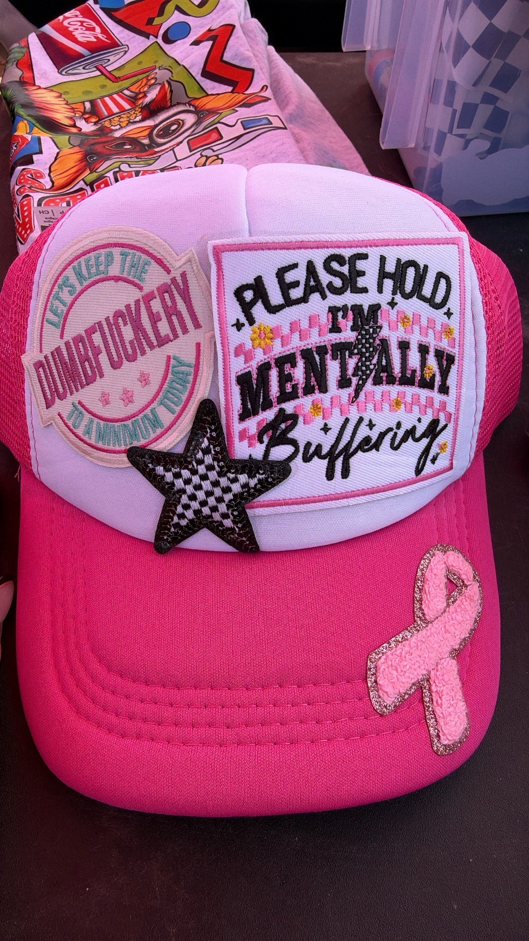 Pink and white trucker hat with slogan patches, a checkered star, and a pink ribbon patch on the brim