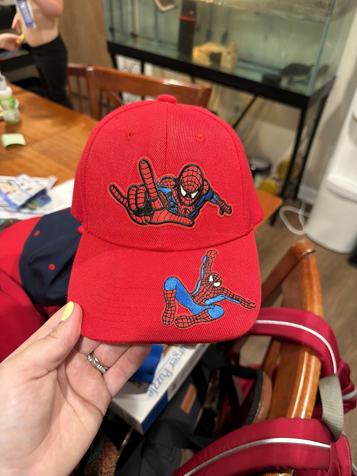 Hand holding a red cap with Spider-Man patches on the front and brim